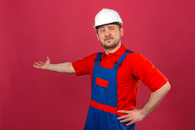 Uniforme da costruzione e casco di sicurezza d'uso dell'uomo del costruttore che indicano da parte con la palma aperta della mano che mostra a copia spazio sorridendo sicuro sopra la parete rosa isolata