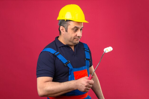 Uniforme da costruzione e casco di sicurezza d'uso dell'uomo del costruttore che stanno con il rullo di pittura e che lo esaminano con il fronte serio sopra la parete rosa isolata