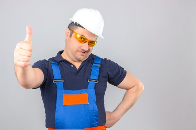 Uniforme da costruzione e casco di sicurezza d'uso dell'uomo del costruttore con il sorriso sul fronte che sbatte le palpebre e che mostra i pollici su sopra la parete bianca isolata
