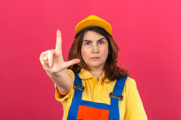 Uniforme della costruzione della donna del costruttore e cappuccio giallo che indicano con l'espressione alta ed arrabbiata del dito che non mostra gesto sopra la parete rosa isolata