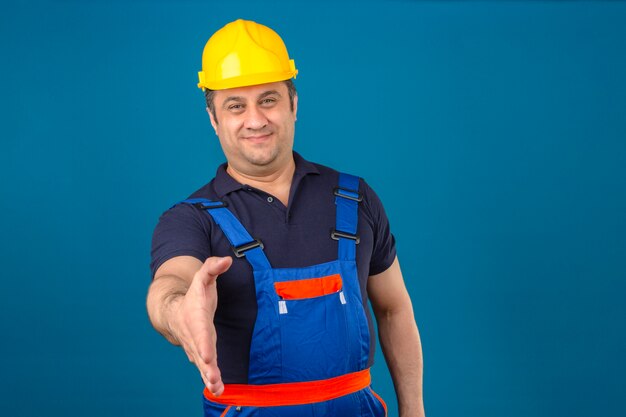 Uniforme di costruzione da portare dell'uomo del costruttore e casco di sicurezza che offrono la mano per stringere saluto e accogliere gesto sopra la parete blu isolata