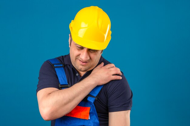 Uniforme di costruzione di mezza età uomo che indossa l'uniforme della costruzione e la spalla commovente del casco di sicurezza che ha dolore sopra la parete blu isolata