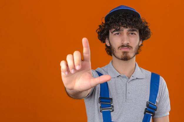 Uniforme e cappuccio d'uso della costruzione del giovane costruttore bello barbuto che indicano con il dito su ed espressione arrabbiata che non mostra gesto sopra la parete arancio isolata