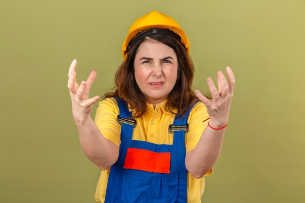 Uniforme edile d'uso del casco della donna della costruzione del costruttore e condizione pazza con l'espressione aggressiva e le armi alzate sopra la parete verde isolata