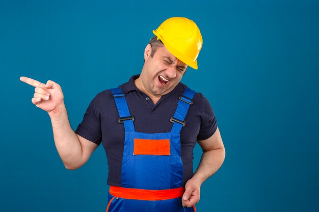 Uniforme edile d'uso del costruttore dell'uomo e casco di sicurezza che indicano con il dito qualcuno e che ridono molto sopra la parete blu isolata