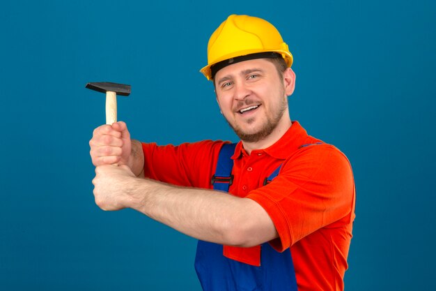 Uniforme edile da portare dell'uomo del costruttore e casco di sicurezza che tengono martello in mani che sorridono allegramente controllando parete blu isolata