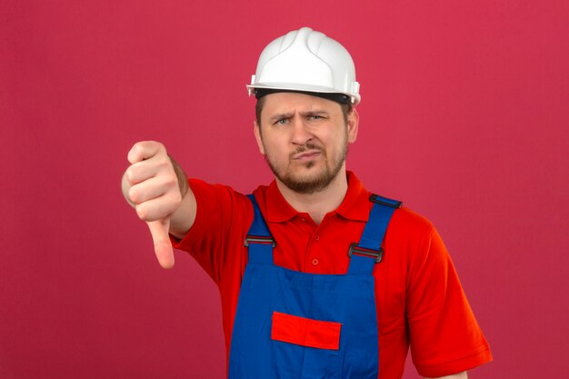 Uniforme edile da portare dell'uomo del costruttore e casco di sicurezza dispiaciuti mostrando pollice giù che controlla parete rosa isolata