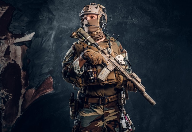 Unità d'élite, soldato delle forze speciali in uniforme mimetica in posa con fucile d'assalto. Foto dello studio contro una parete strutturata scura