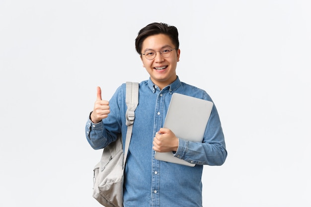 Università, studio all'estero e concetto di stile di vita. Soddisfatto studente maschio asiatico felice in occhiali e camicia che mostra il pollice in su in segno di approvazione, gli piace studiare al college, tenere in mano laptop e zaino.