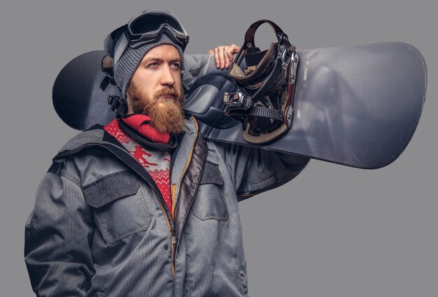 Uno snowboarder rosso brutale con la barba piena in un cappello invernale e occhiali protettivi vestito con un cappotto da snowboard in posa con lo snowboard in uno studio, guardando lontano. Isolato su sfondo grigio.