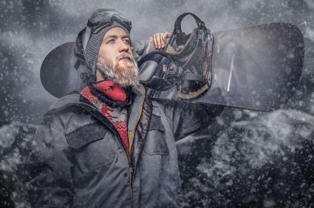 Uno snowboarder rosso brutale con la barba piena in un cappello invernale e occhiali protettivi vestito con un cappotto da snowboard in posa con lo snowboard sullo sfondo delle montagne.