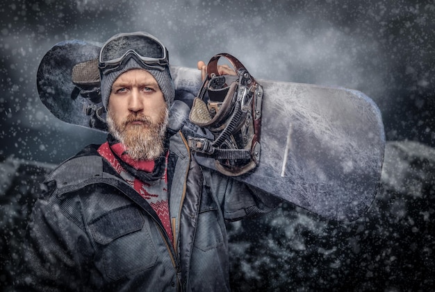 Uno snowboarder rosso brutale con la barba piena in un cappello invernale e occhiali protettivi vestito con un cappotto da snowboard in posa con uno snowboard sullo sfondo delle montagne, guardando una telecamera.