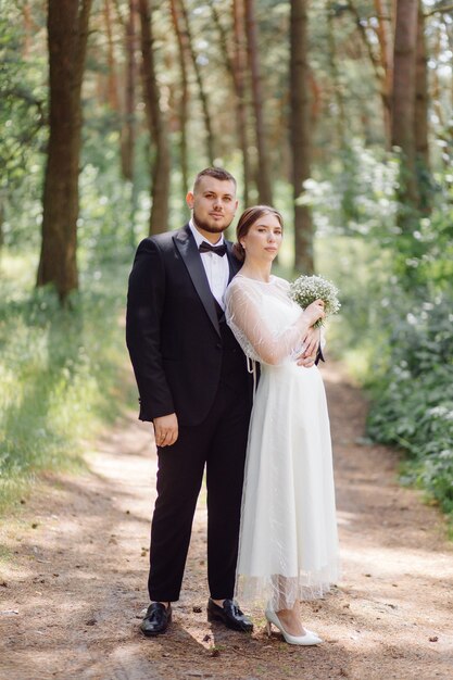 Uno sposo barbuto ed elegante in abito e una bella sposa bionda in abito bianco con un bouquet in mano sono in piedi e si abbracciano nella natura nella foresta di pini.
