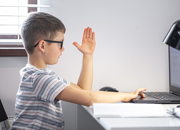 Uno studente di scuola elementare con gli occhiali si siede a un tavolo con un laptop, impara a distanza, alza la mano in una lezione online.