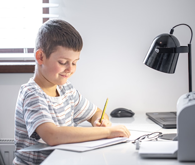 Uno studente di scuola elementare si siede a un tavolo con una lampada e scrive qualcosa su un taccuino.