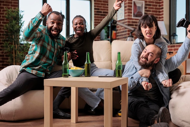 Uomini e donne sorridenti che vincono la competizione dei videogiochi, celebrano la vittoria sulla console TV e si divertono con le bottiglie di birra. Gruppo di persone felici della vittoria del gioco in casa.