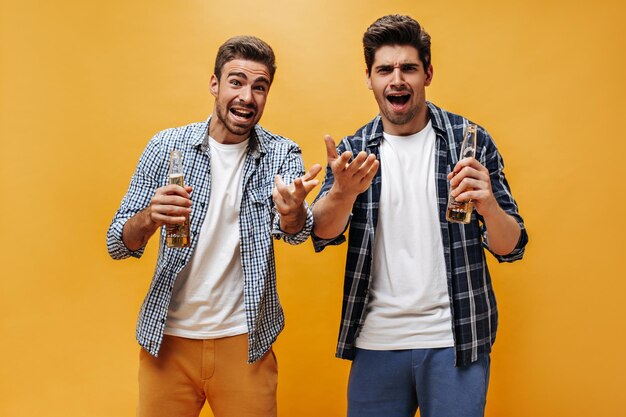 Uomini emotivi delusi in camicie a scacchi pantaloncini colorati e magliette bianche discutono di qualcosa e tengono le bottiglie di birra su sfondo arancione
