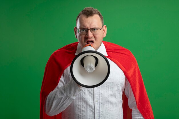 Uomo adulto accigliato del supereroe in mantello rosso con gli occhiali guardando la parte anteriore parlando da altoparlante isolato sulla parete verde