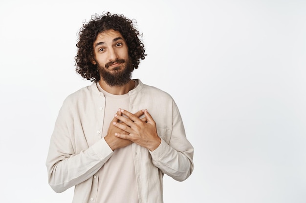 Uomo adulto carino e premuroso che si tiene per mano sul cuore che sembra commosso e lusingato grato per qualcosa in piedi su sfondo bianco