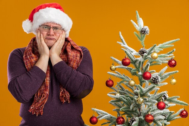 Uomo adulto confuso che indossa occhiali e cappello da Babbo Natale con sciarpa intorno al collo in piedi vicino all'albero di Natale decorato che tiene le mani sul viso isolato sul muro arancione