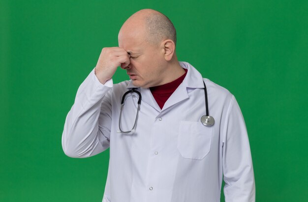 Uomo adulto deluso in uniforme da medico con stetoscopio che si tiene il naso