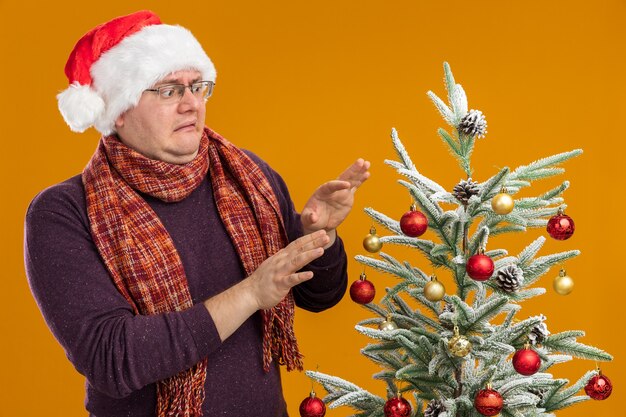 Uomo adulto dispiaciuto con gli occhiali e cappello da Babbo Natale con sciarpa intorno al collo in piedi vicino all'albero di Natale decorato guardando facendo gesto di rifiuto isolato su sfondo arancione