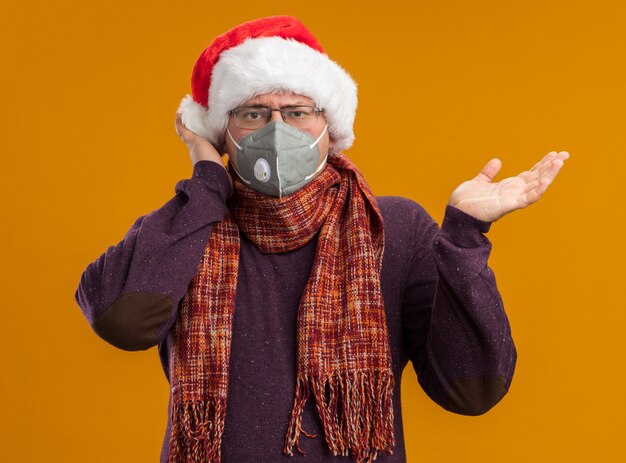uomo adulto impressionato che indossa occhiali maschera protettiva e cappello da Babbo Natale con sciarpa intorno al collo guardando la telecamera che mostra la mano vuota toccando la testa isolata su sfondo arancione
