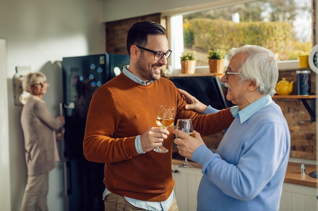 Uomo adulto metà allegro che beve vino con suo padre anziano in cucina