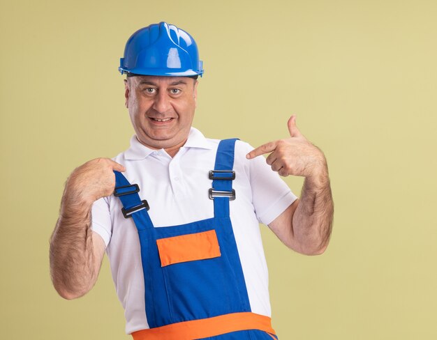 Uomo adulto soddisfatto del costruttore in uniforme punti a se stesso isolato sulla parete verde oliva