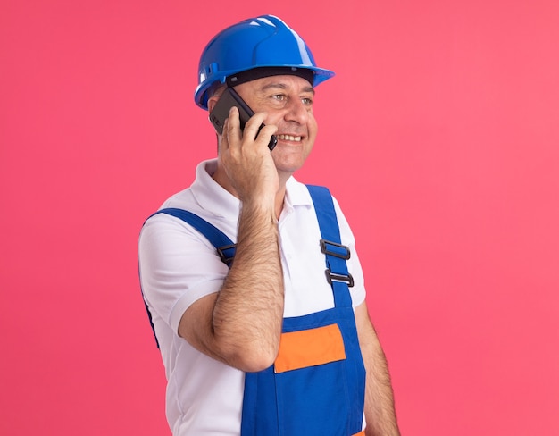 Uomo adulto sorridente del costruttore in colloqui uniformi sul telefono che esamina il lato isolato sulla parete rosa