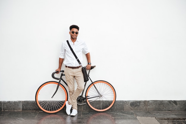 Uomo africano con la bicicletta che sta sulla parete bianca