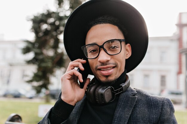 Uomo africano dagli occhi scuri in posa durante la conversazione telefonica. Foto di primo piano all'aperto del ragazzo nero emotivo seduto nel parco con lo smartphone.