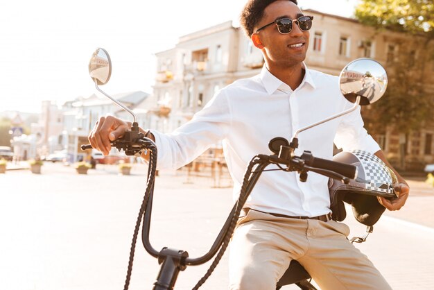 Uomo africano felice che si siede sulla motocicletta moderna all'aperto e distogliere lo sguardo