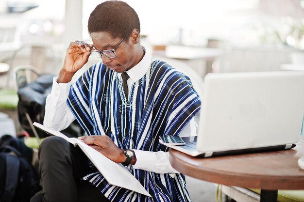 Uomo africano in abiti tradizionali e occhiali seduto dietro il computer portatile al caffè all'aperto e scrivere qualcosa sul suo taccuino