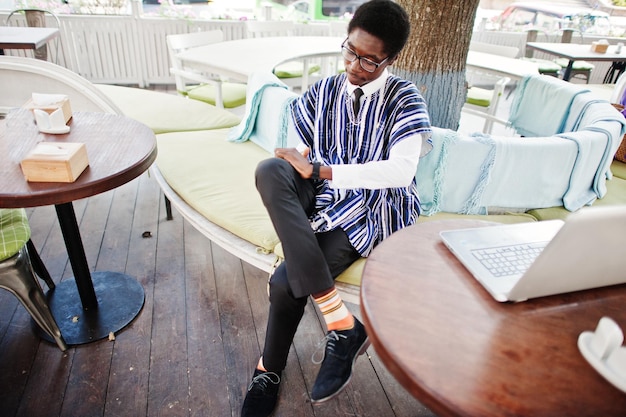 Uomo africano in abiti tradizionali e occhiali seduto dietro il computer portatile al caffè all'aperto