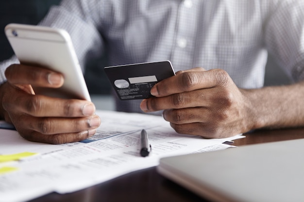 Uomo africano in camicia che paga per merci su internet tramite carta di credito e telefono cellulare