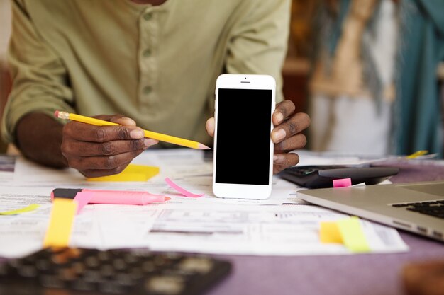 Uomo africano irriconoscibile che tiene la matita gialla, indicandolo allo smart phone dello schermo in bianco nella sua mano