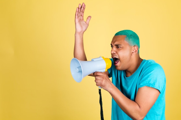 Uomo africano nero in casual sui capelli blu muro giallo che grida gridando nel megafono a sinistra