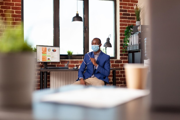 Uomo afroamericano che indossa una maschera e seduto in ufficio per lavorare sulla strategia. Imprenditore aziendale che si prepara a lavorare sulla pianificazione del progetto durante la pandemia di coronavirus.