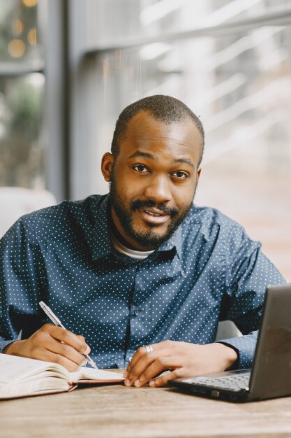 Uomo afroamericano che lavora dietro un computer portatile e scrive su un taccuino. Uomo con la barba seduto in un caffè.