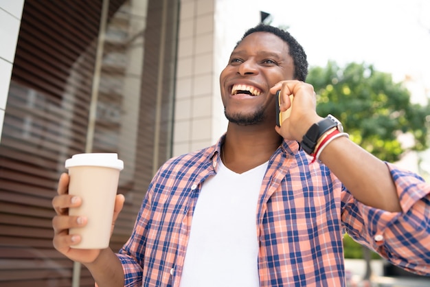 Uomo afroamericano che tiene una tazza di caffè e parla al telefono mentre si cammina per strada.