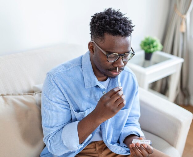 Uomo afroamericano che usa un batuffolo di cotone mentre fa il test PCR del coronavirus a casa uomo che usa il test diagnostico rapido del coronavirus Giovane uomo a casa che usa un tampone nasale per COVID19