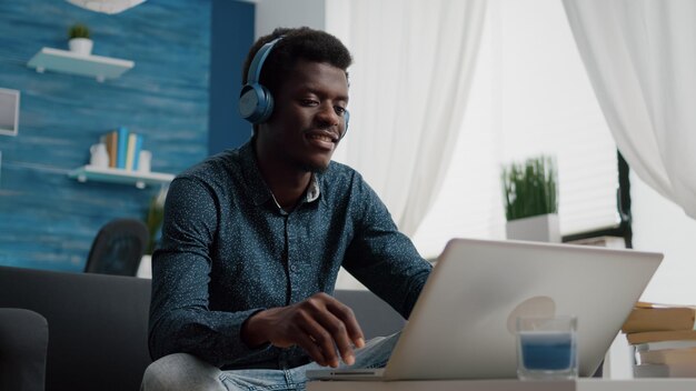 Uomo afroamericano con cuffie per guardare film su servizi di streaming in un luminoso soggiorno, utente di computer concentrato sul lavoro d'ufficio o intrattenimento domestico, sorridente