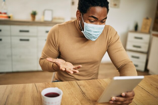 Uomo afroamericano con maschera facciale che utilizza il touchpad mentre effettua una videochiamata a casa