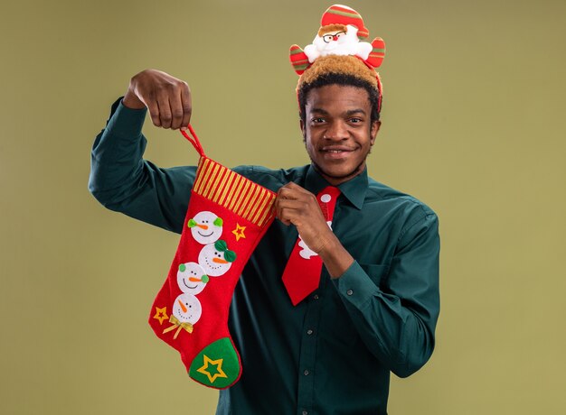 Uomo afroamericano con orlo divertente della santa e cravatta rossa che tiene la calza di Natale che guarda l'obbiettivo sorridente felice e positivo che sta sopra fondo verde
