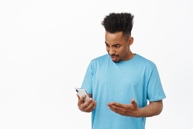 Uomo afroamericano confuso che fissa il suo telefono, guarda lo schermo dello smartphone perplesso e infastidito, si lamenta dell'applicazione, in piedi su bianco