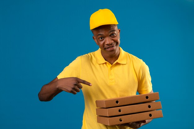 Uomo afroamericano di consegna in camicia di polo gialla e cappuccio che tiene le scatole della pizza e che indica con il dito a loro sorridente in piedi amichevole sul blu isolato