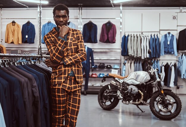 Uomo afroamericano elegantemente vestito che posa con la mano sul mento mentre si trova in un negozio di abbigliamento maschile classico, guardando la fotocamera.