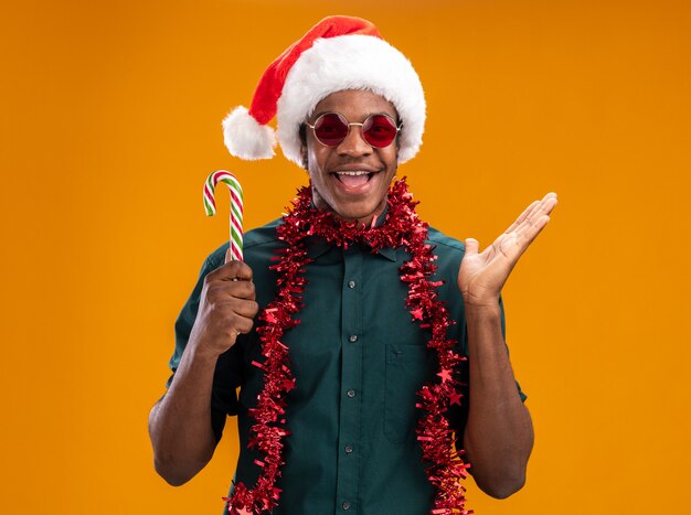 Uomo afroamericano felice in cappello della Santa con la ghirlanda con gli occhiali che tiene il bastoncino di zucchero che sorride allegramente con il braccio alzato che sta sopra la parete arancione