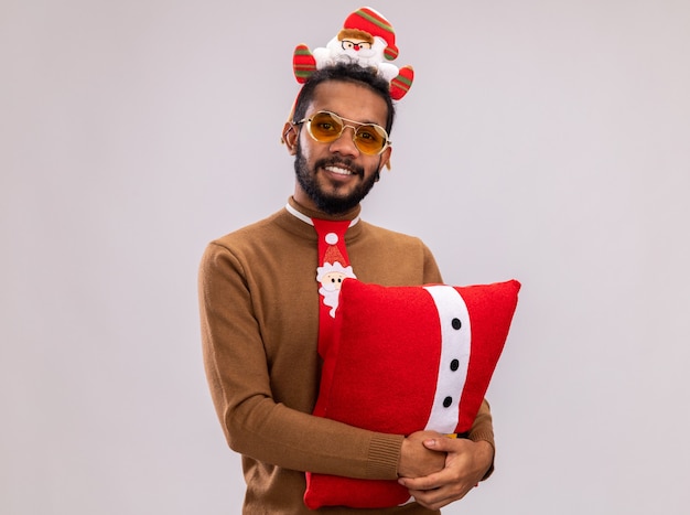 Uomo afroamericano felice in maglione marrone e orlo di santa sulla testa con cravatta rossa divertente che tiene il cuscino di natale che guarda l'obbiettivo con il sorriso sul viso in piedi su sfondo bianco